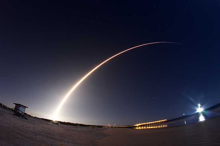 A long-exposure photog of a rocket launch. The rocket's trajectory is depicted as a bright, curved streak of light against the dark night sky.